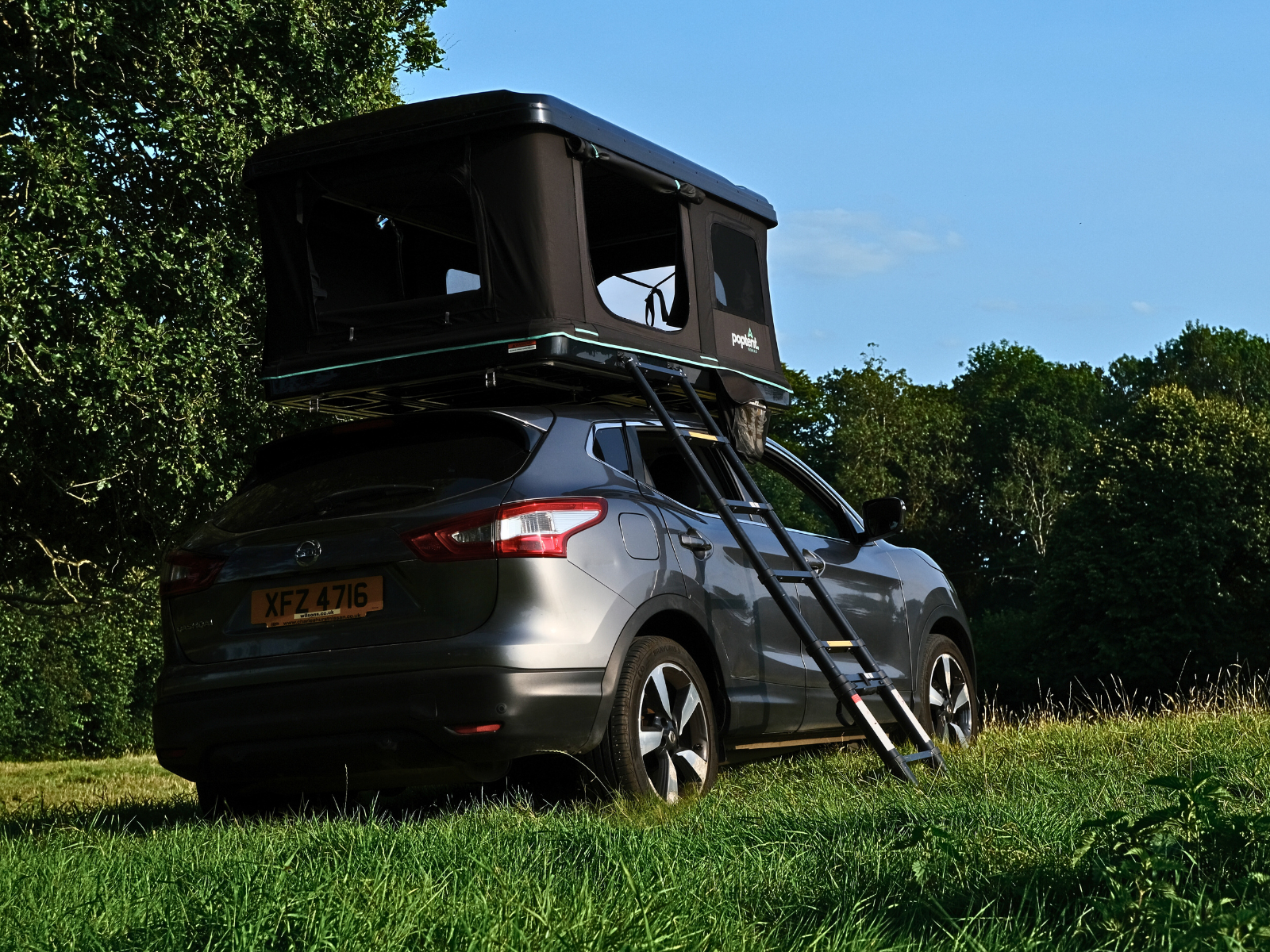 rooftop tent mounted on a grey Nissan Qashqai with ladder extended, parked on grass beside a woodland area under a clear sky
