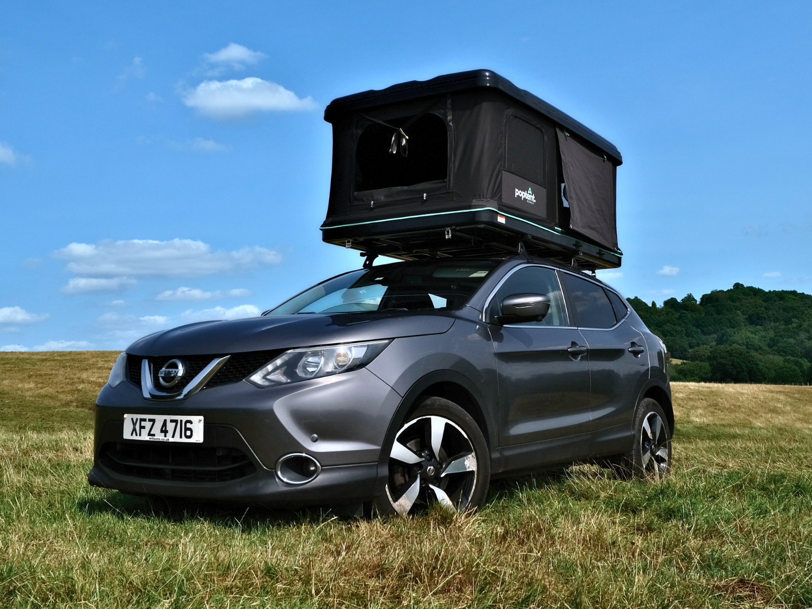 rooftop tent mounted on a grey Nissan Qashqai parked in a grassy field under blue skies