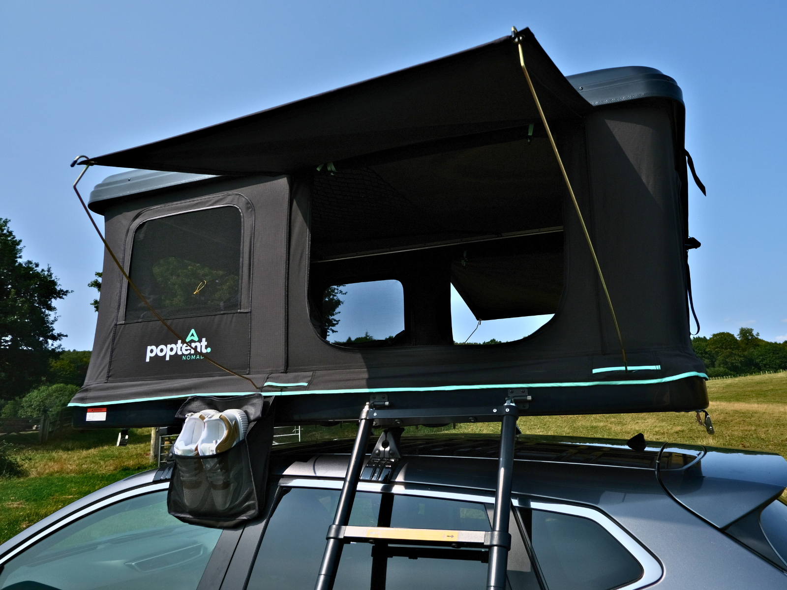 Close-up of Onnio Poptent Nomad rooftop tent mounted on a grey SUV, showing open windows, extended awning, ladder, and exterior shoe pocket