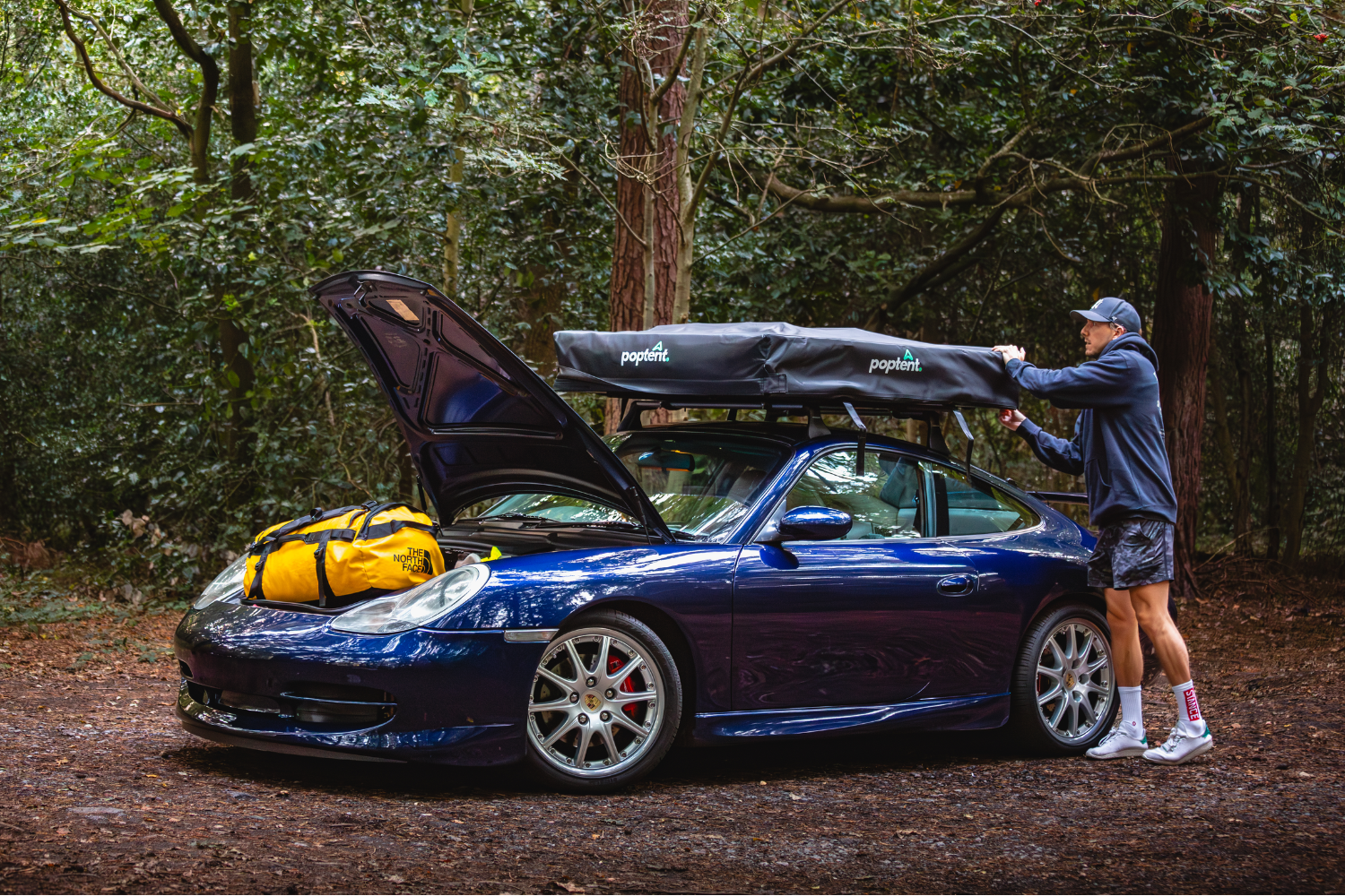 Man attaching a rooftop tent to a blue Porsche 911 parked in a forest, with a yellow North Face duffel bag on the bonnet
