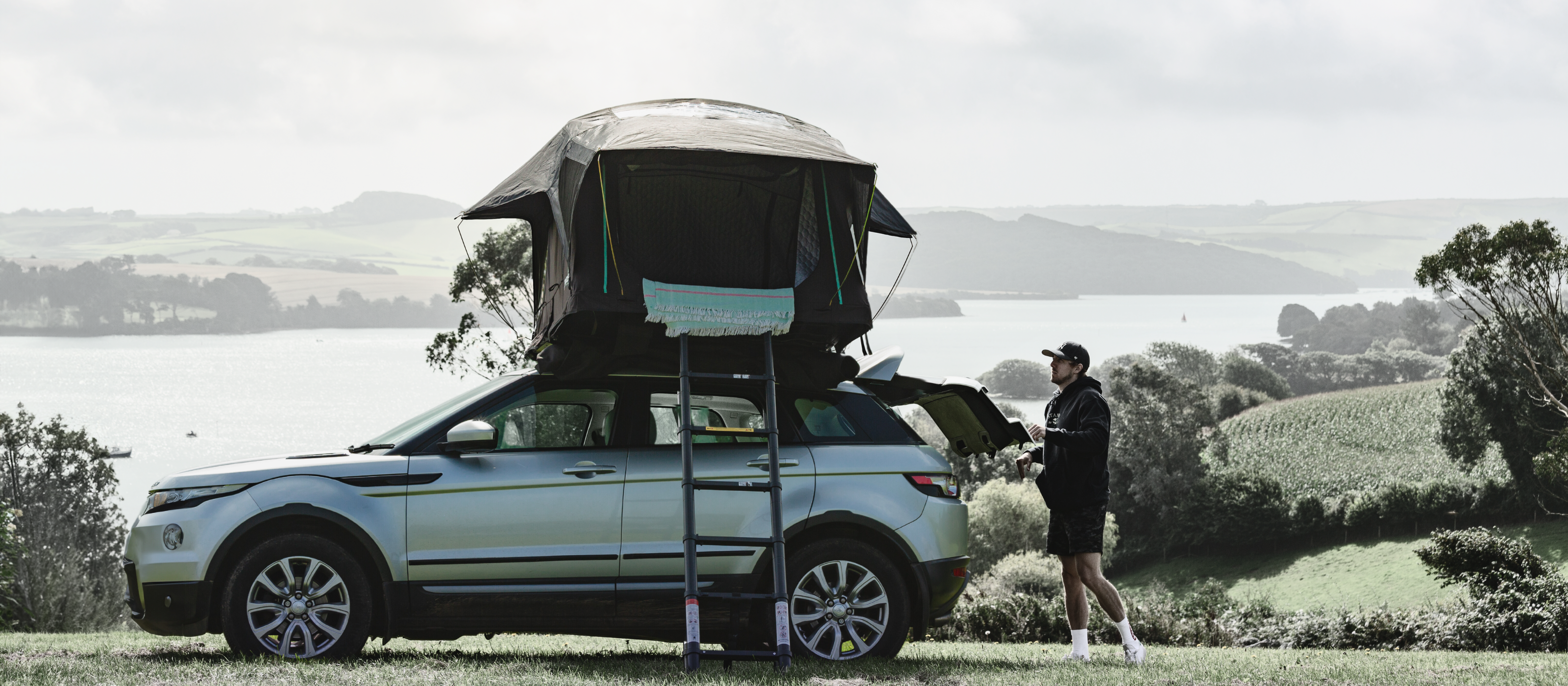 Man standing beside a silver Range Rover with a rooftop tent setup overlooking a scenic lakeside landscape in the countryside