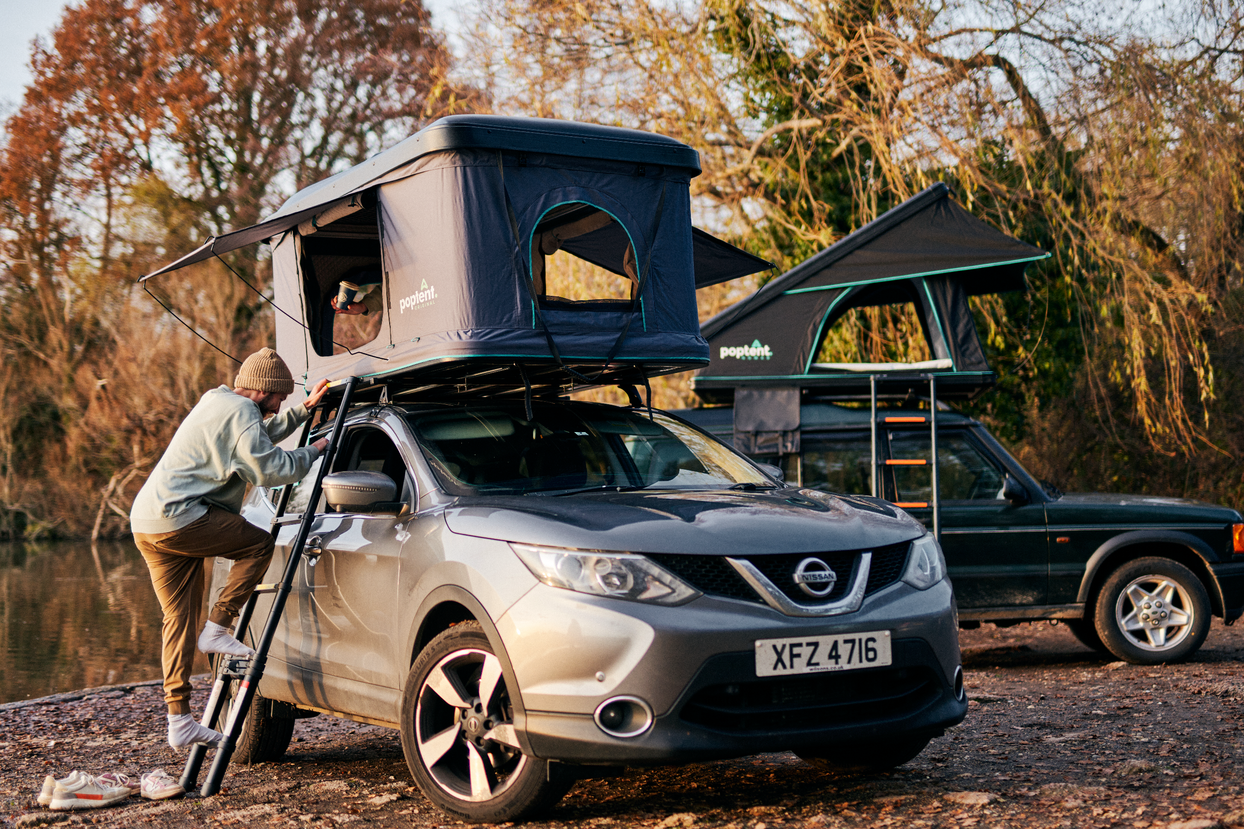 Man climbing into a rooftop tent mounted on a grey Nissan SUV at a lakeside campsite, with another 4x4 and roof tent setup in the background